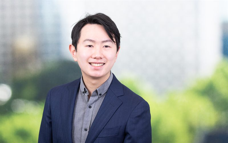 Thomas Ho, smiling in a navy blazer, Melbourne skyline behind